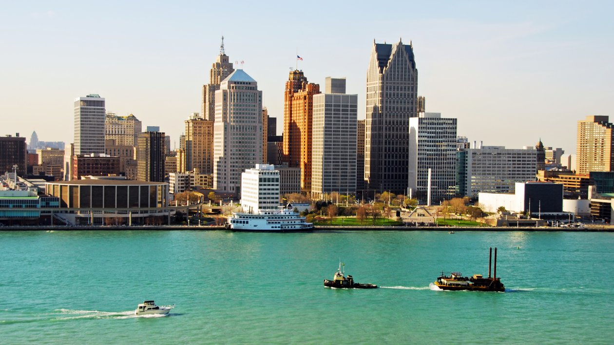 Boats in the Detroit River are seen in the foreground with the Detroit riverfront and skyline in the background, on a sunny summer day.