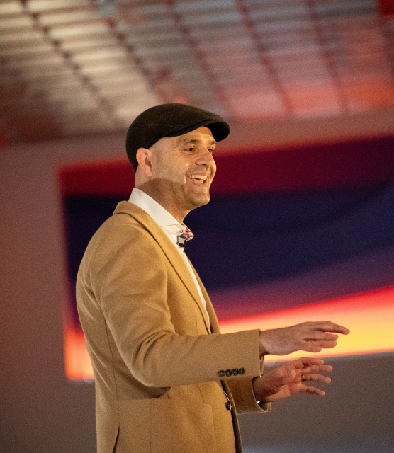 Nikola Danaylov speaks on stage while wearing a newsboy hat at an event at the JRT agency on lighted stage with bright lights in the background