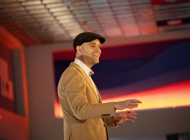 Nikola Danaylov speaks on stage while wearing a newsboy hat at an event at the JRT agency on lighted stage with bright lights in the background